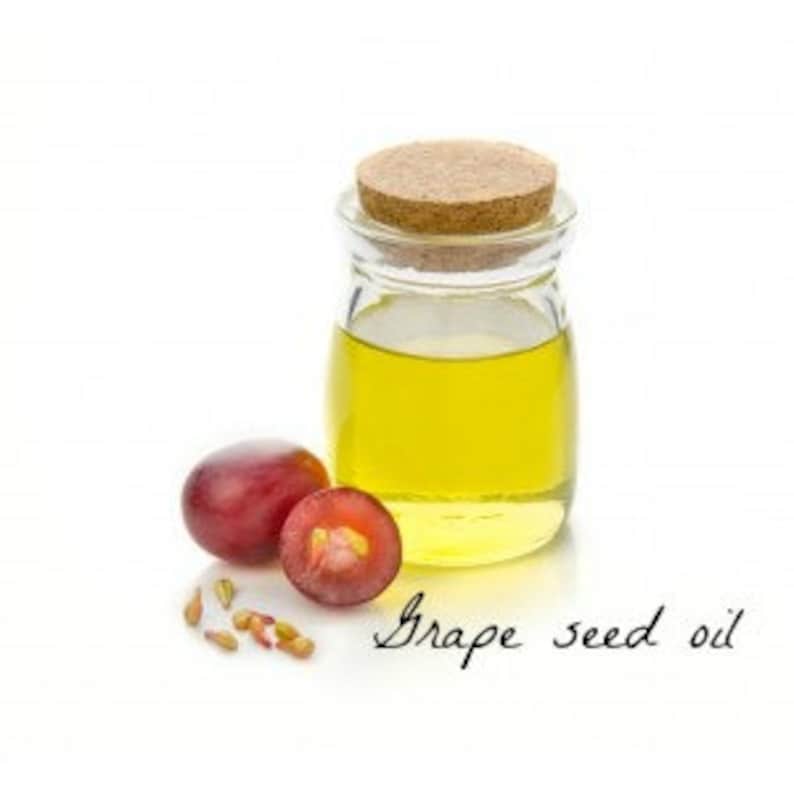 May include: A glass bottle with a cork stopper filled with yellow grape seed oil. Two halved red grapes and grape seeds are next to the bottle. The text "Grape seed oil" is written in cursive below the bottle.