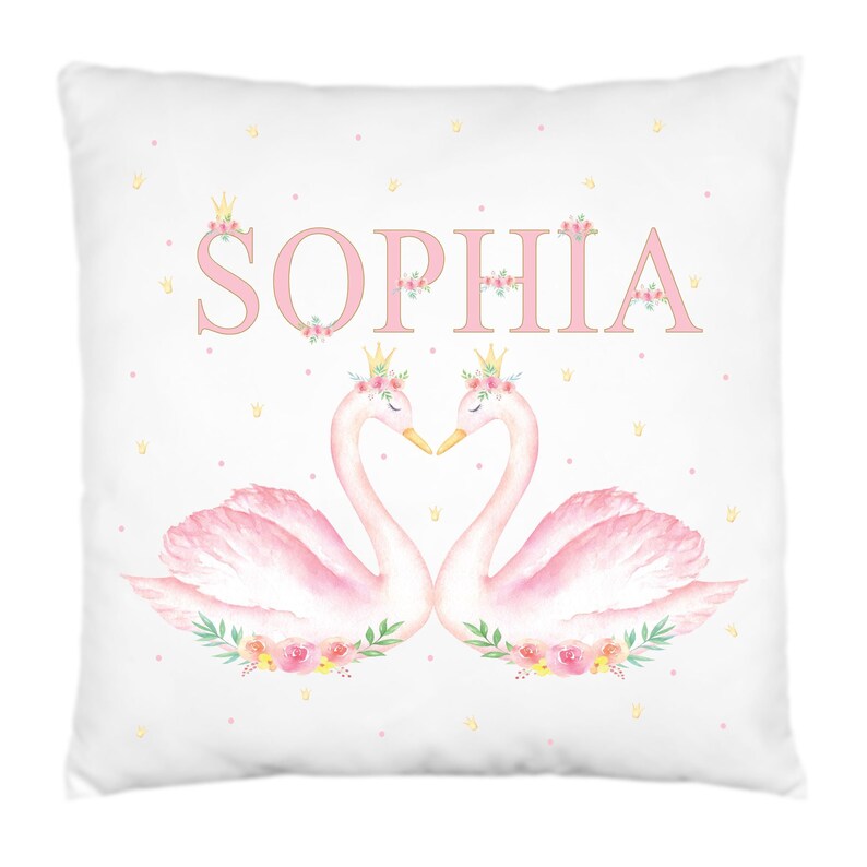 Op de afbeelding: Een wit kussen met roze en gouden accenten met twee roze zwanen met bloemenkronen en de naam "SOPHIA" in roze letters.
