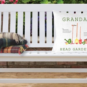 May include: White wooden bench with a cushion that says "Grandad Head Gardener" with a gardening theme. The cushion has a green, red, and yellow colour scheme with a bird, watering can, shovel, rake, and gardening gloves. There are two plaid blankets on the bench.