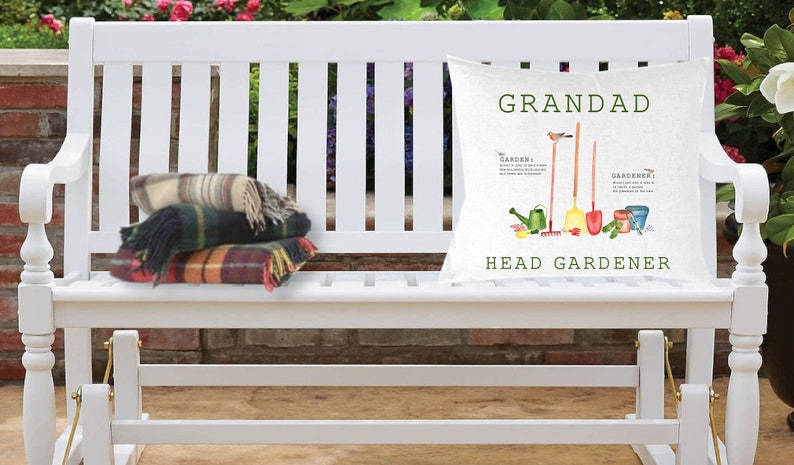 May include: White wooden bench with a cushion that says "Grandad Head Gardener" with a gardening theme. The cushion has a green, red, and yellow colour scheme with a bird, watering can, shovel, rake, and gardening gloves. There are two plaid blankets on the bench.