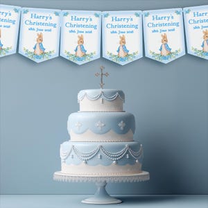 K&ouml;nnte beinhalten: Eine dreist&ouml;ckige, hellblaue Torte mit wei&szlig;er Glasur und Perlenakzenten, gekr&ouml;nt von einem silbernen Kreuz. Dar&uuml;ber ein Banner mit dem Text "Harry's Christening 28th June 2036" und einer Peter Hase-Illustration.