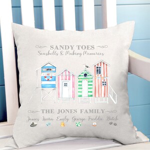 May include: A light grey throw pillow with a watercolour illustration of beach huts and the text "SANDY TOES Seashells & Making Memories THE JONES FAMILY James Laura Emily George Freddie Butch".