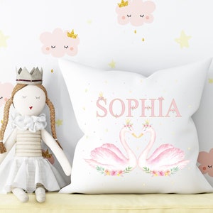 Op de afbeelding: Een wit kussen met een roze en wit zwanenontwerp en de naam "Sophia" in roze gedrukt. Het kussen is versierd met gouden kronen en bloemen.