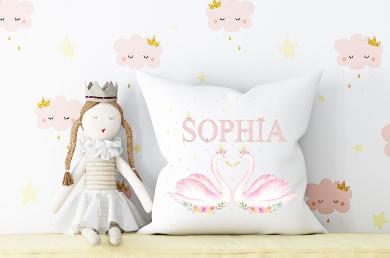 Op de afbeelding: Een wit kussen met een roze en wit zwanenontwerp en de naam "Sophia" in roze gedrukt. Het kussen is versierd met gouden kronen en bloemen.