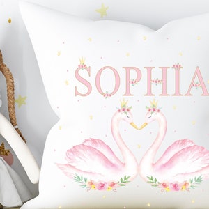 Op de afbeelding: Een wit kussen met roze zwanen en bloemmotieven. De naam "SOPHIA" is geschreven in roze letters met gouden kronen boven elke letter.