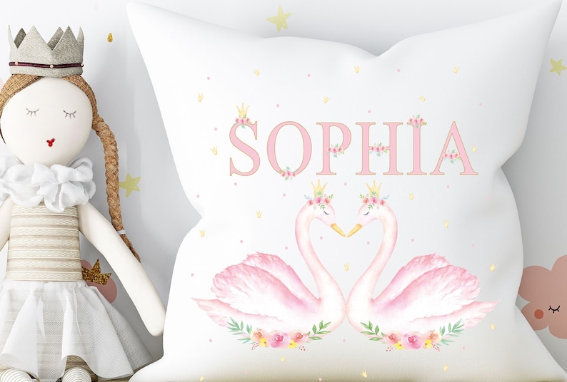 Op de afbeelding: Een wit kussen met roze zwanen en bloemmotieven. De naam "SOPHIA" is geschreven in roze letters met gouden kronen boven elke letter.