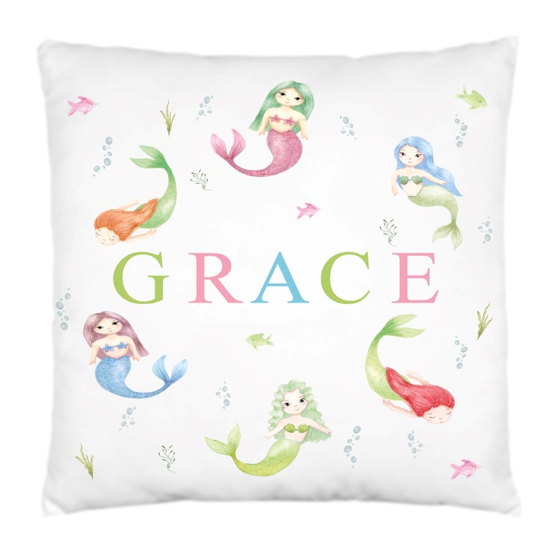 May include: White pillow with a watercolor design of mermaids and the name "GRACE" in colorful letters. The mermaids are in a circle around the name, with their tails pointing outwards. The background is white with a light blue, green, and pink watercolor design.