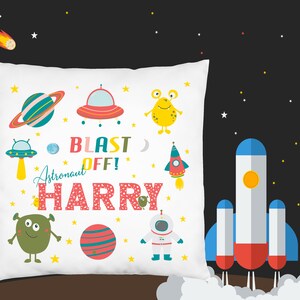 K&ouml;nnte beinhalten: Wei&szlig;es Kissen mit einem Weltraum-Motiv. Es zeigt eine Rakete, Planeten, Au&szlig;erirdische und den Text "Blast Off! Astronaut Harry".