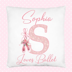 K&ouml;nnte beinhalten: Ein wei&szlig;es Kissen mit rosa Schriftzug "Sophia Loves Ballet". Der Buchstabe "S" ist gro&szlig; und mit einem rosa Blumenmuster verziert. Ein Paar rosa Ballettschuhe mit einer rosa Schleife und floralen Akzenten befinden sich links vom Buchstaben "S".