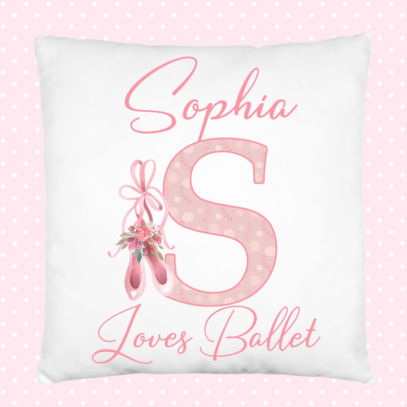K&ouml;nnte beinhalten: Ein wei&szlig;es Kissen mit rosa Schriftzug "Sophia Loves Ballet". Der Buchstabe "S" ist gro&szlig; und mit einem rosa Blumenmuster verziert. Ein Paar rosa Ballettschuhe mit einer rosa Schleife und floralen Akzenten befinden sich links vom Buchstaben "S".