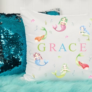May include: White pillow with a watercolor design of mermaids and the name "GRACE" in colorful letters. The pillow is surrounded by a teal sequin pillow and a light blue fuzzy surface.