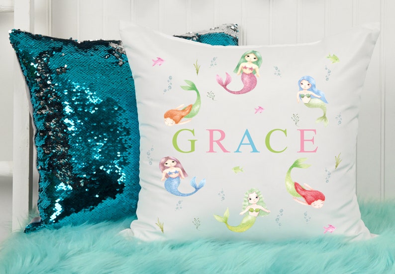 May include: White pillow with a watercolor design of mermaids and the name "GRACE" in colorful letters. The pillow is surrounded by a teal sequin pillow and a light blue fuzzy surface.