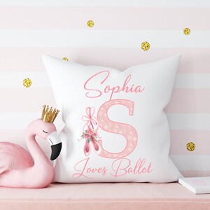 K&ouml;nnte beinhalten: Ein wei&szlig;es Kissen mit einem rosafarbenen, gepunkteten Buchstaben S und rosafarbenen Ballettschuhen mit Blumen. Das Kissen tr&auml;gt den Namen Sophia und den Text "Loves Ballet".
