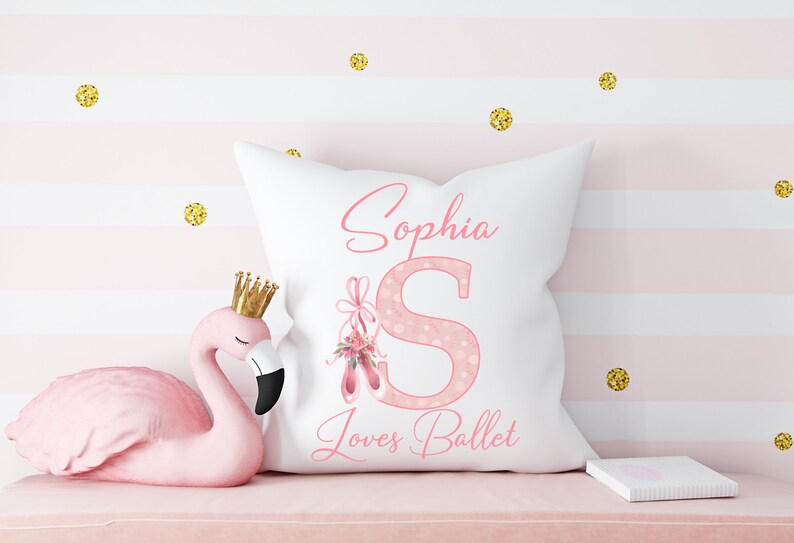 K&ouml;nnte beinhalten: Ein wei&szlig;es Kissen mit einem rosafarbenen, gepunkteten Buchstaben S und rosafarbenen Ballettschuhen mit Blumen. Das Kissen tr&auml;gt den Namen Sophia und den Text "Loves Ballet".