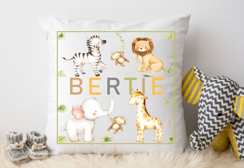 K&ouml;nnte beinhalten: Ein wei&szlig;es Kissen mit gr&uuml;nem Rand, auf dem ein Zebra, ein L&ouml;we, ein Elefant und eine Giraffe mit dem Namen "BERTIE" in braunen, gelben und grauen Buchstaben abgebildet sind.
