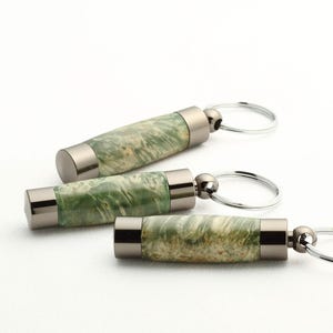 Puede incluir: Tres llaveros con cuerpos cilíndricos de madera verde y crema y detalles metálicos plateados. Cada llavero tiene un anillo plateado adjunto. Los llaveros están dispuestos en diagonal sobre un fondo blanco.