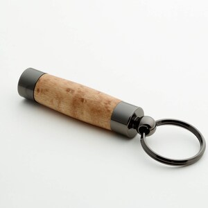 Può includere: Un portachiavi cilindrico con un corpo centrale in legno marrone chiaro caratterizzato da una venatura naturale, rifinito alle estremità con inserti in metallo grigio scuro lucido e collegato a un anello metallico circolare abbinato.