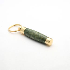 Könnte beinhalten: Ein grüner und goldener Schlüsselanhänger mit einem Schlüsselring und einem zylindrischen Holzkörper mit einer goldenen Kappe.