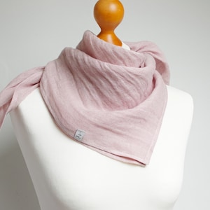 May include: A dusty pink triangular scarf with a crinkled texture draped around a mannequin neck. The scarf features a small, rectangular grey fabric tag on the lower edge with the letter Z printed in black, showcasing a simple, lightweight design.