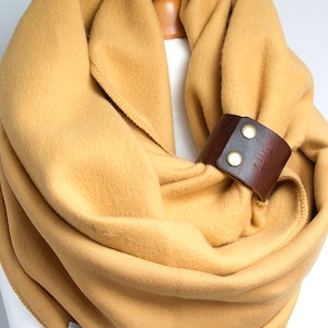 Könnte beinhalten: Senfgelber Infinity-Schal mit einer dunkelbraunen Leder-Manschette, die mit zwei messingfarbenen Druckknöpfen befestigt ist. Der Schal besteht aus einem weichen, flauschigen Material, ideal für kaltes Wetter. Die Leder-Manschette verleiht dem Accessoire einen rustikalen Touch.