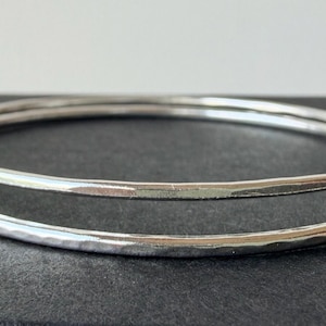 May include: Two thin, circular silver bangles with a hammered, reflective surface texture. The bracelets are stacked slightly offset, showcasing the metallic sheen and the subtle, uneven indentations created by the hammering process on the metal.