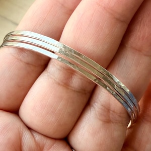 May include: A set of three thin, silver-colored metal bangles with a hammered, textured finish. The bracelets are stacked together, featuring a reflective, metallic surface that catches the light across their rounded, slender bands.