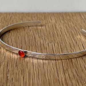 May include: A thin, polished silver-tone metal cuff bracelet featuring a single, small, round, vibrant orange-red cabochon stone set securely into the band. The bracelet has an open-ended design with a smooth, reflective surface finish.