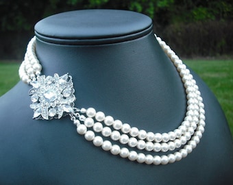 May include: A three-strand pearl necklace with a silver and crystal brooch. The necklace is on a black display stand.