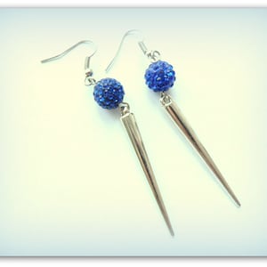 May include: A pair of dangle earrings with a blue rhinestone ball and a silver-coloured, elongated spike. The earrings have a silver hook closure. The earrings are displayed against a light blue background.