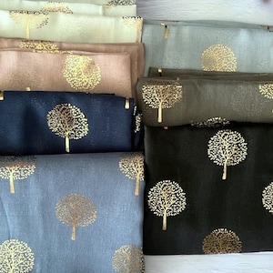 May include: A stack of seven scarves with a gold tree pattern on a light wood surface. The scarves are in various colours, including beige, brown, blue, and grey.