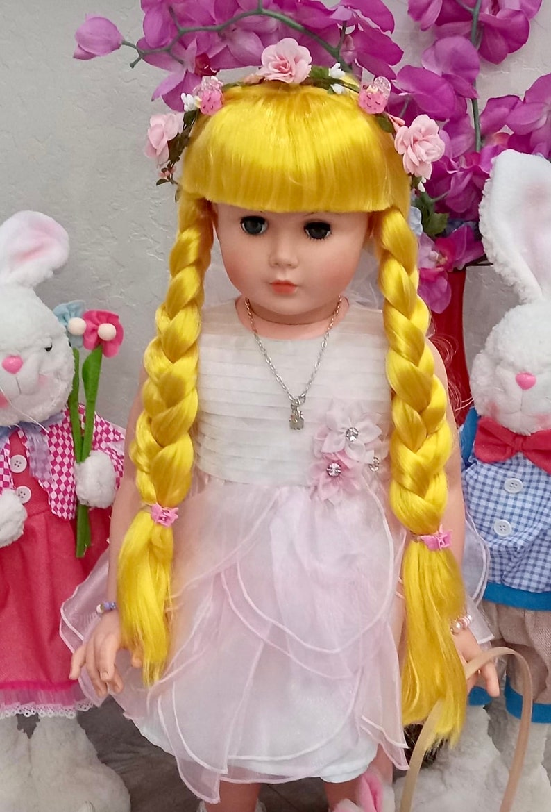 May include: A yellow haired doll wearing a white dress with pink ruffles and a flower crown. The doll has blue eyes and is holding a basket. The doll is standing next to a white bunny with a pink bow.