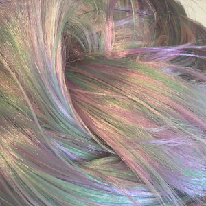 May include: A close-up of iridescent hair extensions in shades of purple, green, and blue. The hair is arranged in a loose, flowing style, showcasing the shimmering colors.