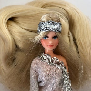 May include: A doll with long blonde hair wearing a silver headband and a silver and white dress.