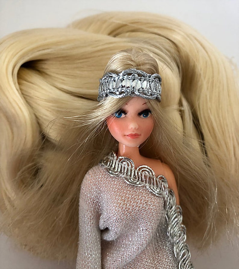 May include: A doll with long blonde hair wearing a silver headband and a silver and white dress.