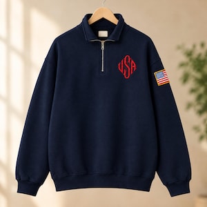 Op de afbeelding: Een marineblauw sweatshirt met een korte rits, voorzien van een rood geborduurd USA-monogram op de linkerborst en een rechthoekige Amerikaanse vlag-patch op de linkermouw. Het kledingstuk heeft een hoge kraag en geribbelde boorden.
