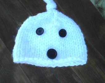 May include: White knitted baby hat with three black buttons for eyes and a mouth.