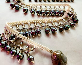 May include: A beaded necklace featuring a cream-colored woven seed bead base with dangling dark iridescent pearls and small faceted clear crystals. The piece terminates in a single large, smooth, mottled brown stone pendant at one end.