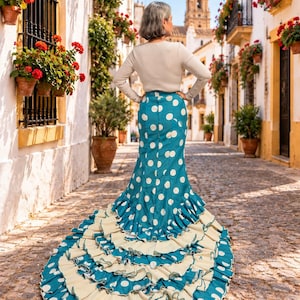 May include: A long-sleeved beige top paired with a floor-length teal skirt featuring large white polka dots. The skirt has a fitted waist and a dramatic, multi-layered ruffled hem that extends into a train across the cobblestone ground.
