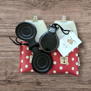 May include: Black castanets with a polished finish, resting on a red polka-dot case. The case has a white interior and a silver clasp. A tag reads "Castanuelas Del Sur."