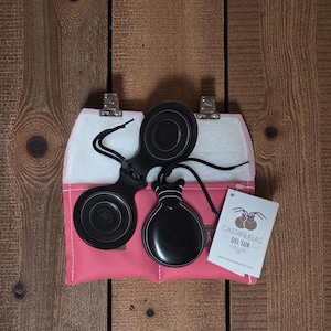 May include: Black castanets with black cords, resting on a pink and white pouch. The castanets are displayed against a wooden background. A tag with the text "CASTANUELAS DEL SUR" is attached to the pouch.
