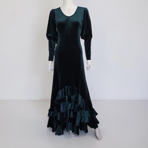May include: A floor-length dark emerald green velvet dress featuring long sleeves with gathered shoulders, a scoop neckline, and a skirt finished with multiple tiers of ruffled fabric at the hemline, displayed on a white mannequin.