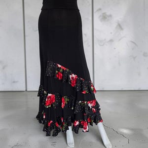 May include: A floor-length black skirt featuring a high-waisted design and a diagonal tiered ruffle hem. The ruffles are decorated with a vibrant red floral pattern and small white polka dots against a black background, creating a layered effect.