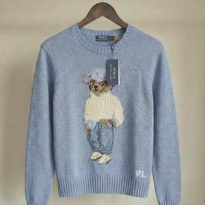 Puede incluir: Un suéter de cuello redondo azul claro con un gráfico tejido de un oso de peluche que lleva un suéter crema, pantalones azules y un sombrero. El suéter tiene cuello, puños y dobladillo acanalados, con las letras RL bordadas en blanco.