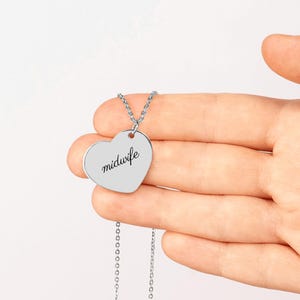 May include: A silver-toned heart-shaped pendant necklace featuring the word midwife engraved in a black cursive script. The pendant is attached to a delicate silver-toned chain link necklace, showcasing a smooth metallic finish.