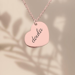 Puede incluir: Un collar con colgante en forma de coraz&oacute;n de color oro rosa que presenta la palabra doula grabada en una tipograf&iacute;a cursiva negra. El colgante cuelga de una delicada cadena a juego en oro rosa, creando una pieza de joyer&iacute;a sencilla.