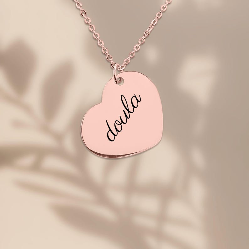 Puede incluir: Un collar con colgante en forma de coraz&oacute;n de color oro rosa que presenta la palabra doula grabada en una tipograf&iacute;a cursiva negra. El colgante cuelga de una delicada cadena a juego en oro rosa, creando una pieza de joyer&iacute;a sencilla.