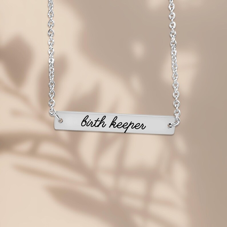 Puede incluir: Un collar de barra horizontal plateada con una cadena delicada y un colgante rectangular. El colgante est&aacute; grabado con el texto "birth keeper" en una fuente cursiva negra, centrado sobre la superficie de metal pulido.