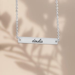 Pode incluir: Um colar de barra horizontal prateada com uma corrente fina e uma placa de metal polido. A palavra "doula" est&aacute; gravada em uma fonte cursiva preta no centro da barra retangular, que est&aacute; presa &agrave; corrente em ambas as extremidades.