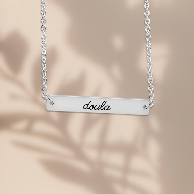 Pode incluir: Um colar de barra horizontal prateada com uma corrente fina e uma placa de metal polido. A palavra "doula" est&aacute; gravada em uma fonte cursiva preta no centro da barra retangular, que est&aacute; presa &agrave; corrente em ambas as extremidades.