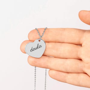 May include: A silver-toned heart-shaped pendant necklace featuring the word doula engraved in a black cursive script. The pendant is attached to a delicate silver-toned chain link necklace, showcasing a smooth, polished metallic finish.